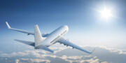 A white commercial airplane flies above the clouds toward a bright sun in a clear blue sky.