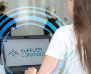 A woman types on a laptop displaying the words Supplier Community with a logo, while two people stand blurred in the background.