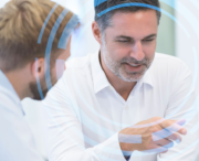 Two men in white shirts have a discussion while looking at something out of frame, with blue circular digital graphics overlaid on the image.