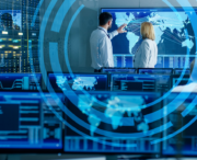 Two people in a control room discuss data on a large world map display, with multiple computer monitors showing global supply chain collaboration and information in the foreground.