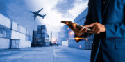 Person in a suit using a tablet at a shipping container yard, with forklifts moving containers and an airplane flying overhead.