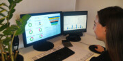 A woman sits at a desk using two monitors displaying data dashboards with charts and graphs, next to a plant and a calculator.