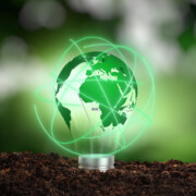 A glass light bulb with a green world map inside sits on soil, surrounded by glowing green lines, symbolizing global sustainable energy.