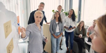 A group of people stands around a whiteboard as a woman points to a diagram, appearing to lead a discussion or presentation in a modern office setting.