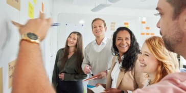 Five people stand by a whiteboard with sticky notes, collaborating and smiling, as one person writes or points at the board.