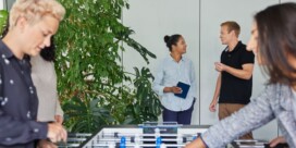 Four people are in an office; two are playing foosball in the foreground, while two others stand and talk near plants in the background.