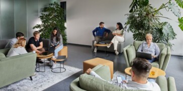 Several people are seated in a modern lounge area, working on laptops, conversing, and using mobile devices, surrounded by green plants and comfortable furniture.