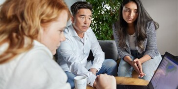 Three people sit around a table, looking intently at a laptop screen; one holds a mug, and there is a leafy green plant in the background.