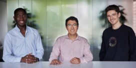 Three men stand and sit behind a white table, smiling at the camera, with a blurred office background behind them.