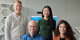 Four people pose indoors in front of a monitor, with two standing and two sitting, dressed in business-casual attire.