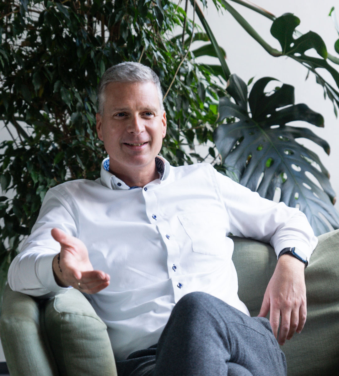 A man with gray hair in a white shirt sits on a green chair, gesturing with one hand. Large green plants in the background create an atmosphere of health and success.