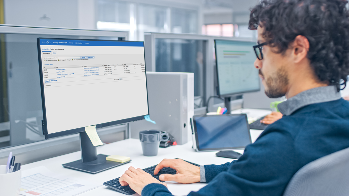 A man with curly hair and glasses works at a desk, viewing a financial or business management dashboard on a desktop computer in a modern office.