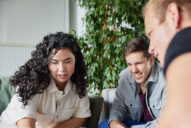 Three people sit close together indoors, engaged in conversation and looking at something out of frame, with a large green plant in the background.