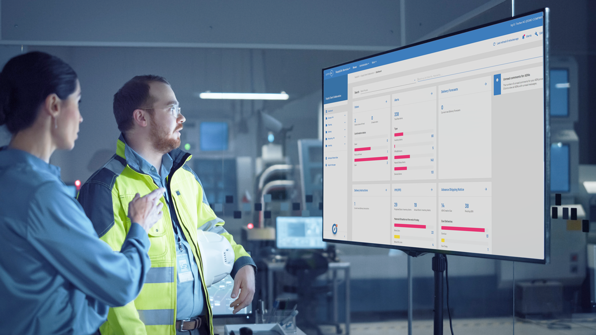 Two people in industrial work attire review data charts and graphs displayed on a large monitor in a modern factory or control room setting.