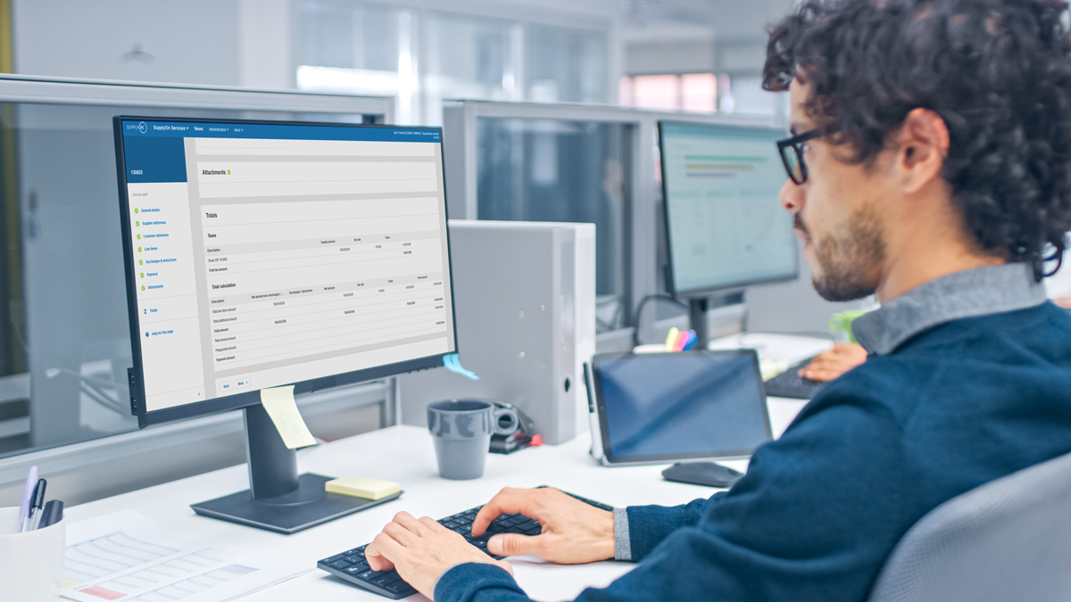 A man sits at a desk in an office, working on a desktop computer displaying a database or management software interface.