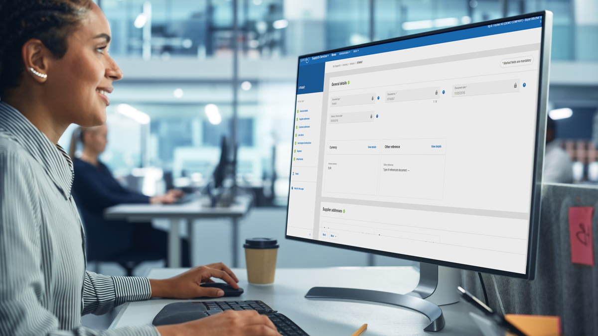 A person sits at a desk in an office, using a desktop computer displaying a business management software interface.