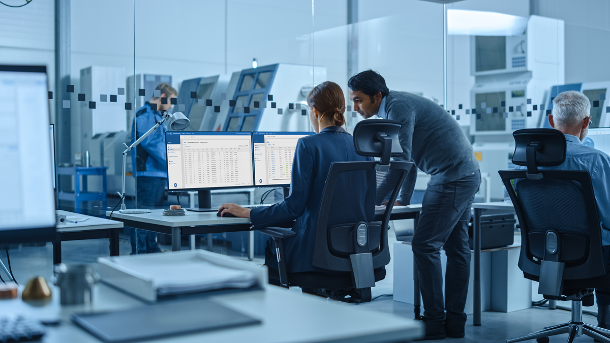 Several people work at computer desks in a modern office with large windows; two colleagues are discussing data displayed on dual monitors.