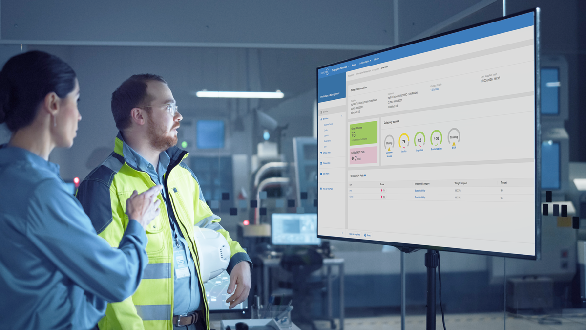 Two people in a modern industrial setting review data and charts displayed on a large monitor, discussing information related to operations or equipment performance.