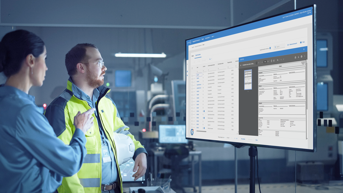 Two people in industrial workwear review spreadsheets and documents displayed on a large monitor in a modern, high-tech workplace.
