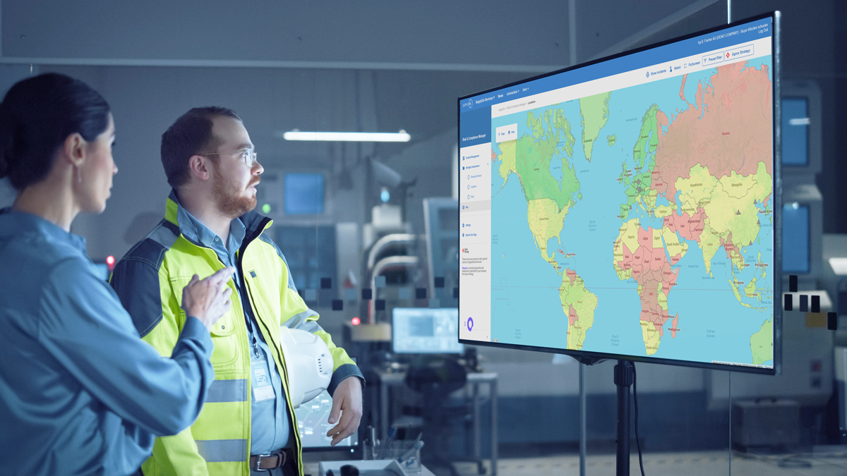 Two people in work attire discuss data displayed on a large monitor showing a colour-coded world map in a modern control room.