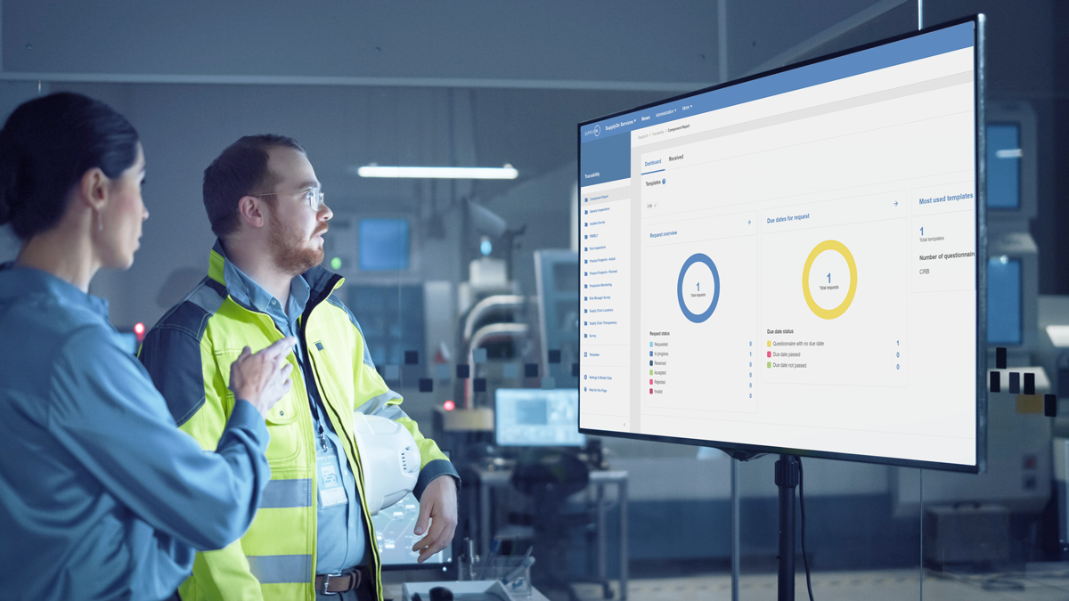 Two people in work attire review data displayed on a large monitor in a modern industrial control room. The screen shows charts and statistics.