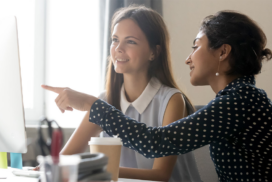 Two women sit at a desk looking at a computer screen. One woman points at the monitor while the other smiles. A coffee cup and office supplies are on the desk.