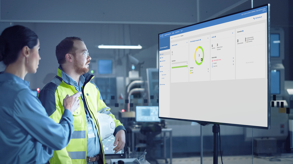 Two people in a modern industrial setting look at a large screen displaying data and charts, possibly discussing performance metrics or operational information.